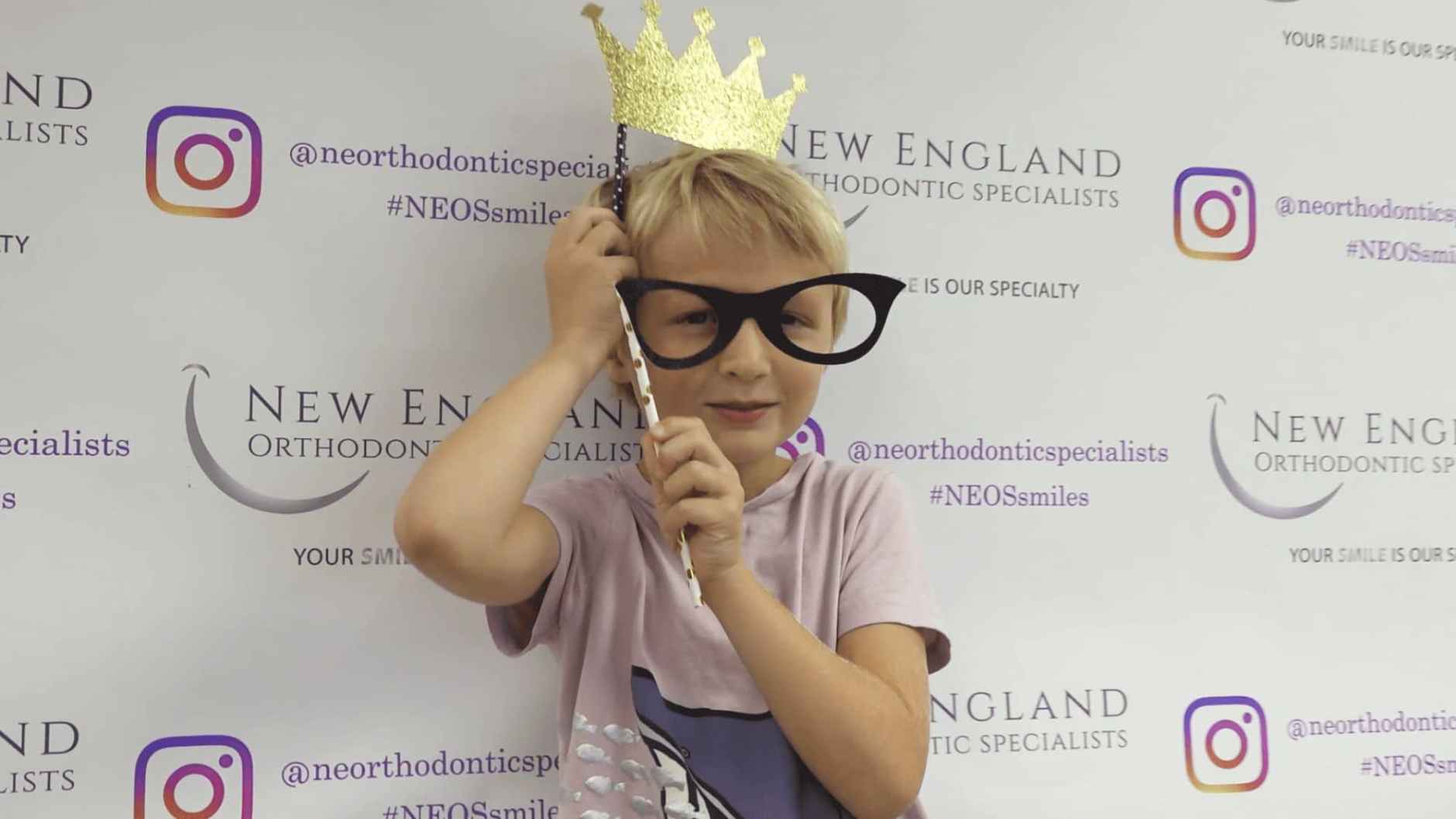 A young boy wearing glasses and a crown stands proudly in front of the New England Orthodontics sign